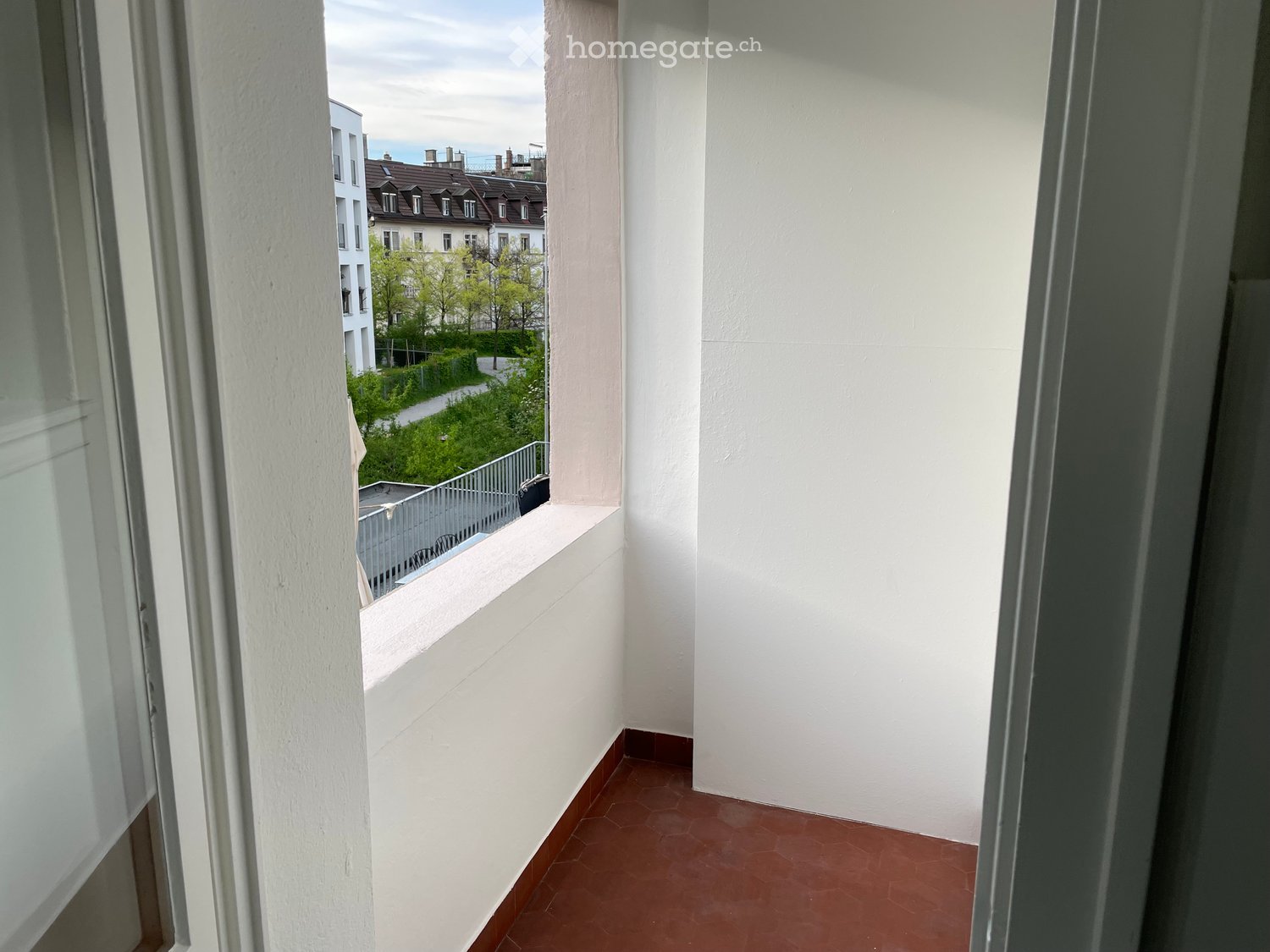 Balcony with red tiled floor, white walls, and a view of a residential area with buildings and greenery.