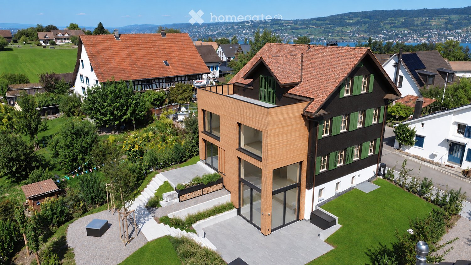 A modern two-story house with a red-tiled roof, large windows, and a balcony overlooking a scenic lake and mountains. The house is surrounded by a well-maintained garden with trees and shrubs.