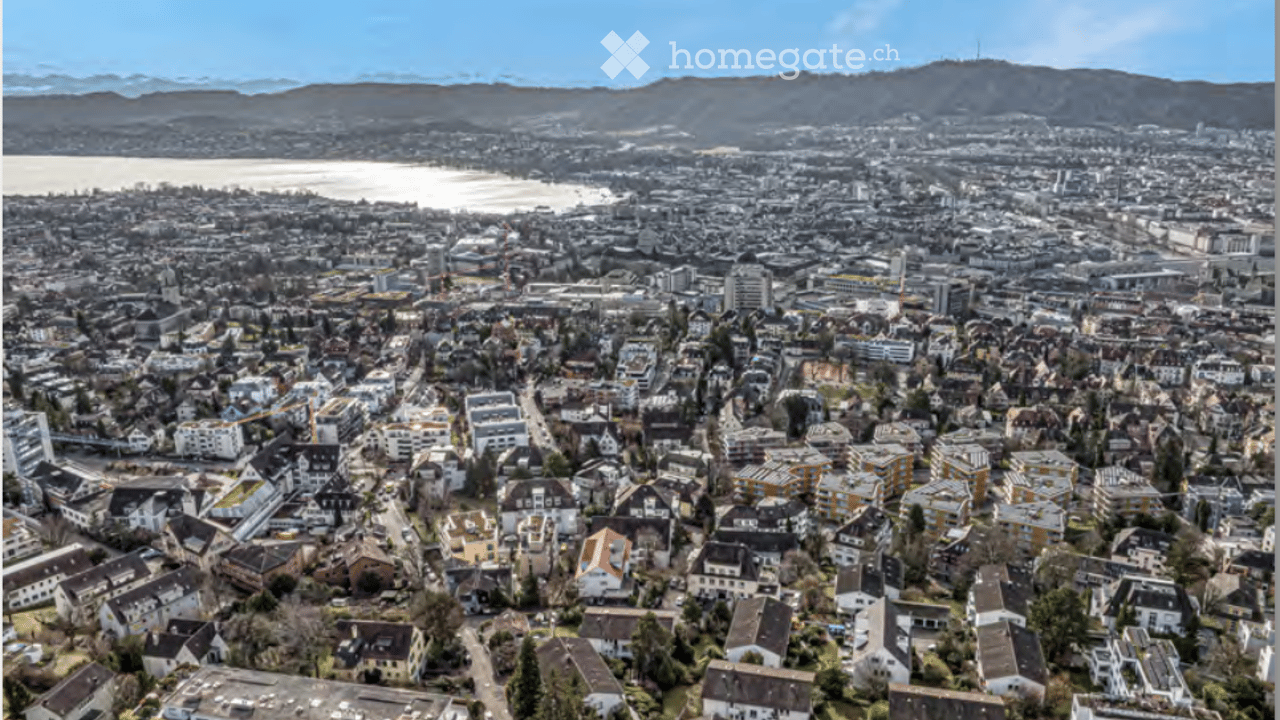 Aerial view of a dense urban area with numerous residential and commercial buildings, surrounded by a body of water and distant mountains.