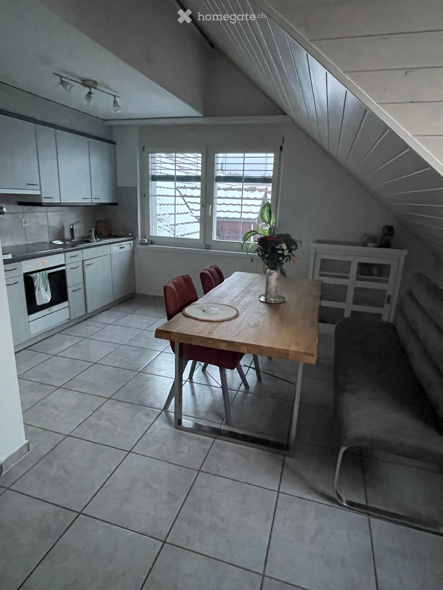 Attic kitchen with white cabinets, tiled floor, wooden dining table with chairs, and a vase with flowers on the table.