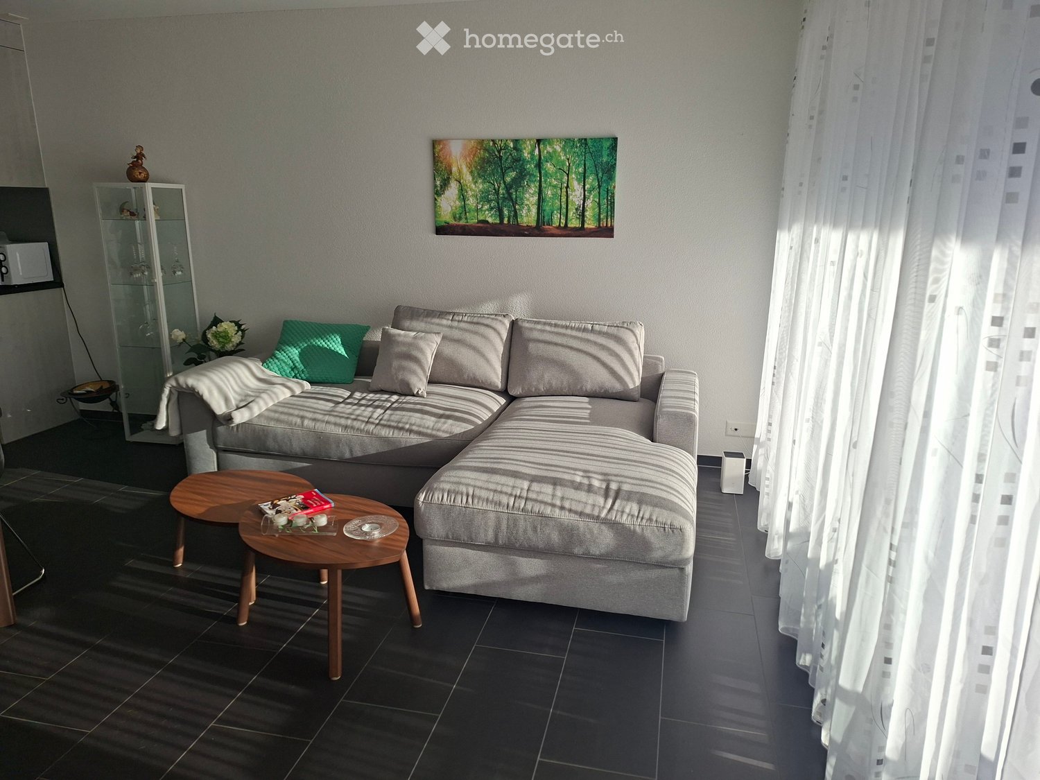 Modern living room with a grey sectional sofa, wooden coffee tables, a large painting, and a glass shelf with decorative items.
