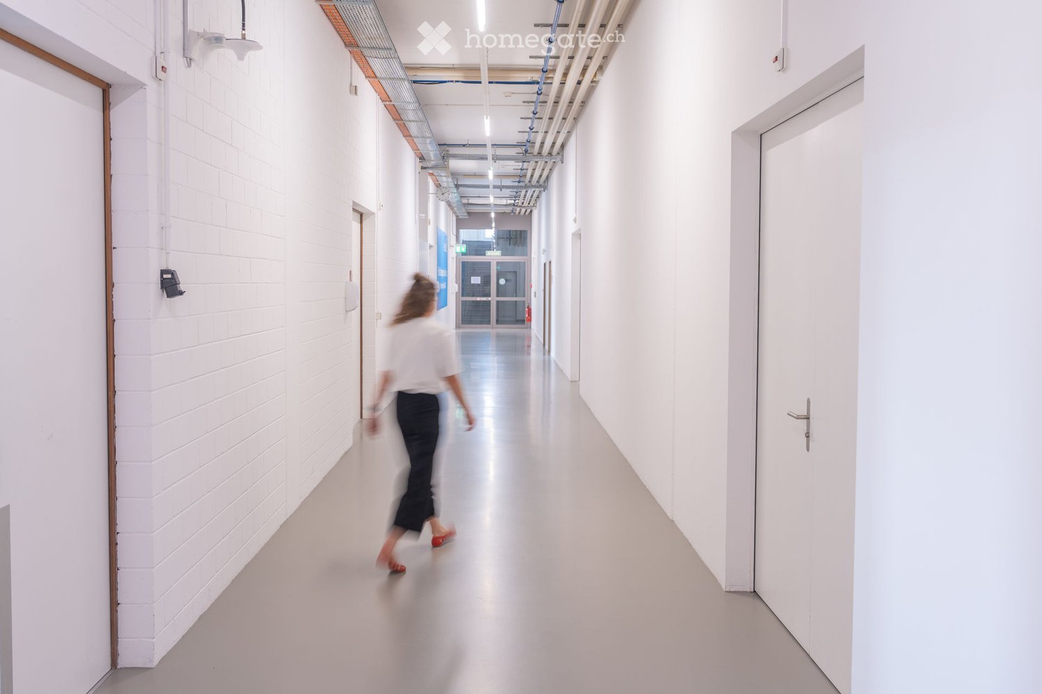 A modern, clean hallway with white walls, a polished concrete floor, and several doors on both sides. Exposed pipes and lighting fixtures are visible on the ceiling.
