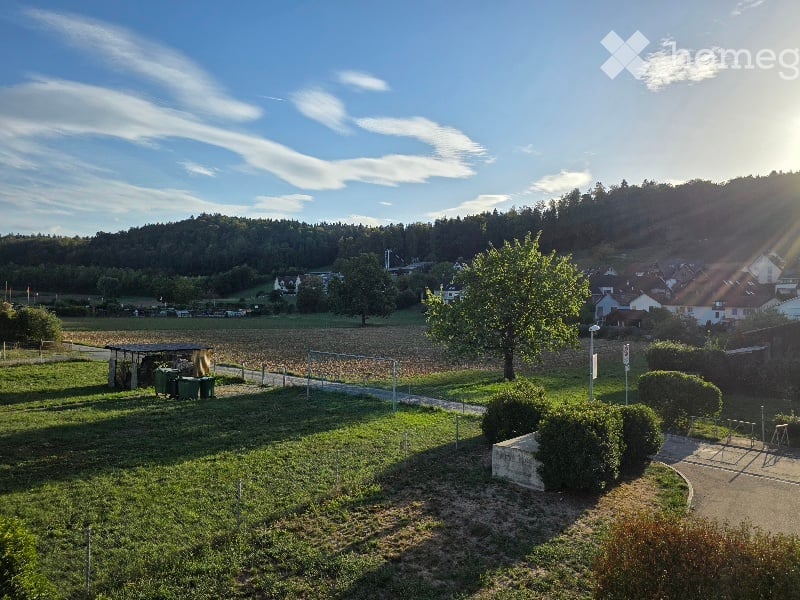A peaceful outdoor area with a grassy lawn, a few trees, and a small structure, surrounded by a rural landscape with houses and hills in the background.