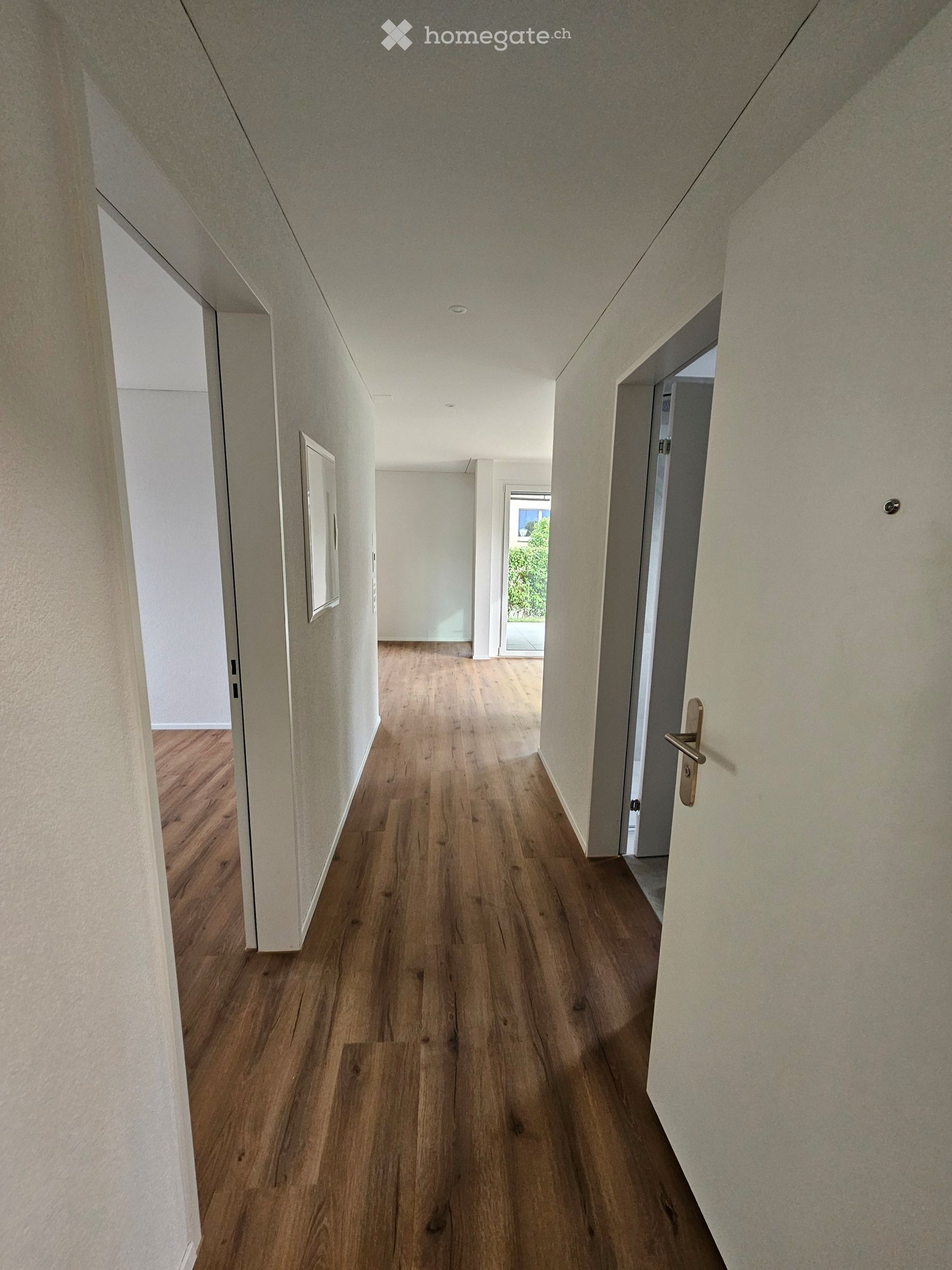 Modern hallway with wooden flooring, white walls, and recessed lighting. Doors and a framed picture are visible on the left side.
