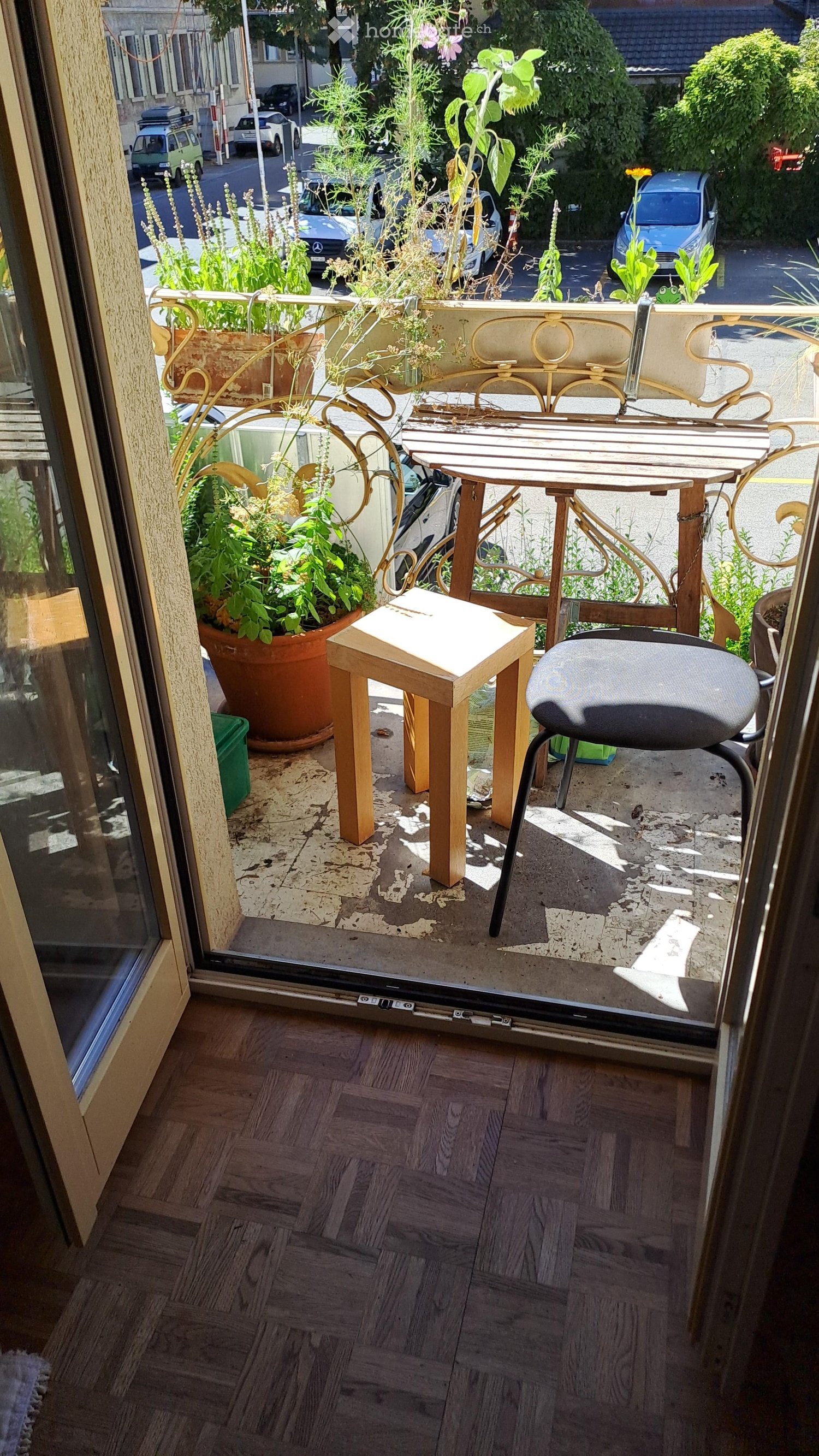 Balcony with wooden table, chair, and potted plants, overlooking a street with parked cars.
