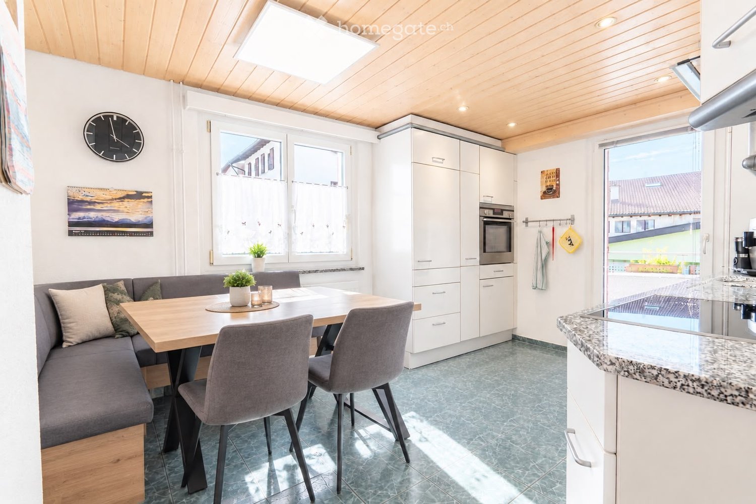Bright, modern kitchen and dining area with white cabinets, a grey sofa, a wooden dining table, and grey chairs. Features include a large window, a clock on the wall, and a potted plant on the table.