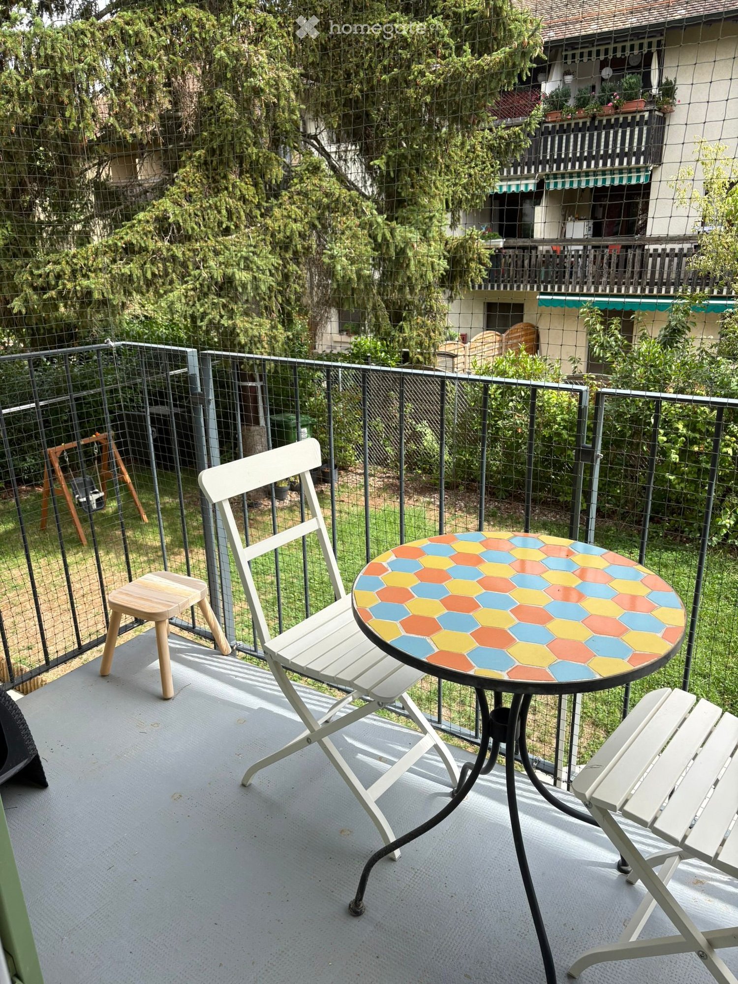 Balcony with a colorful hexagonal table and white folding chairs, surrounded by a metal fence and greenery.