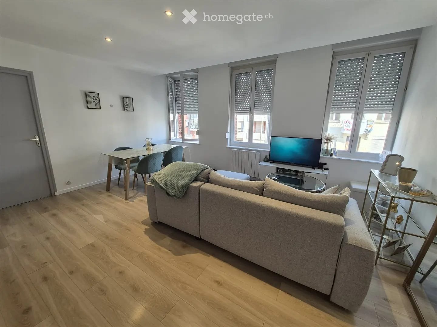 Modern living room with light wooden flooring, large windows, a grey sofa, coffee table, dining area with chairs, and a TV stand.