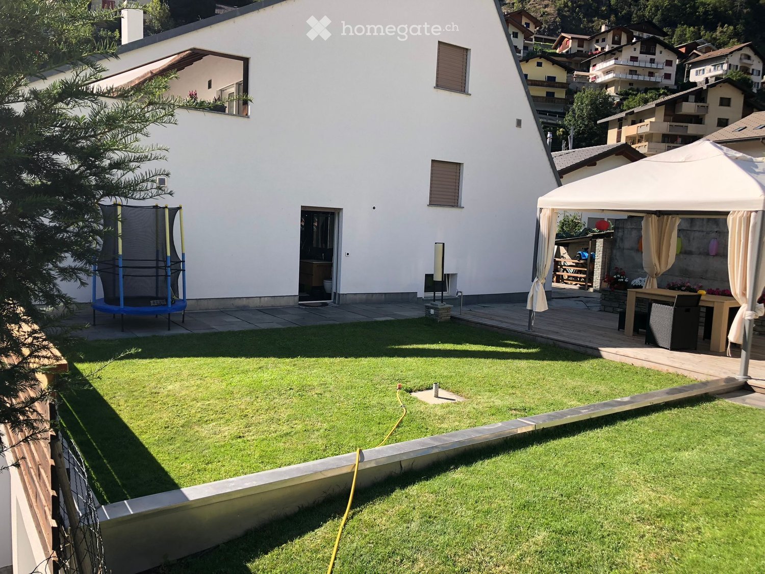 Well-maintained garden with a trampoline, patio area with a table and chairs, and a covered outdoor seating area with curtains.