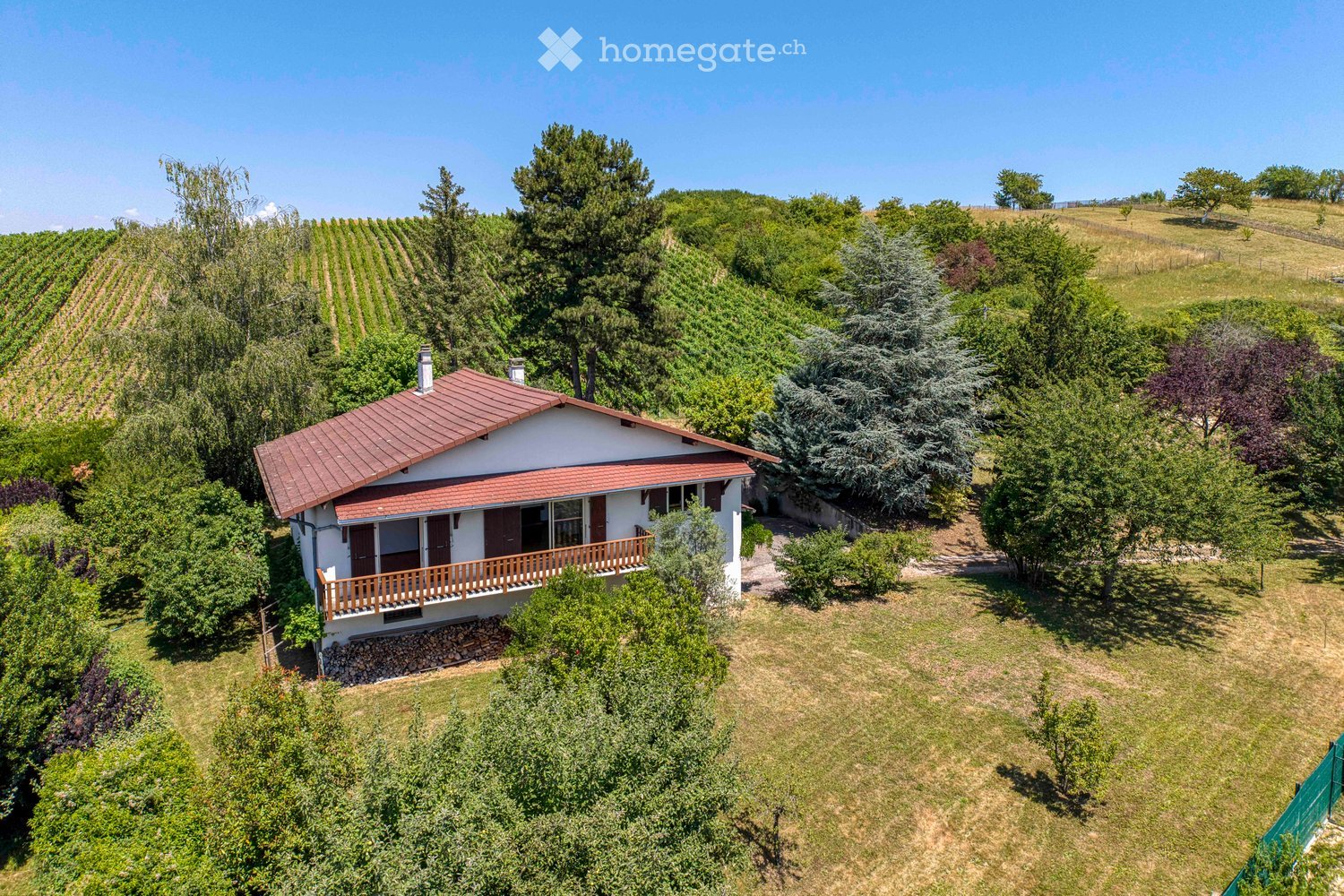 A two-story house with a red-tiled roof, white exterior, and a wooden balcony overlooking a vineyard and lush greenery.