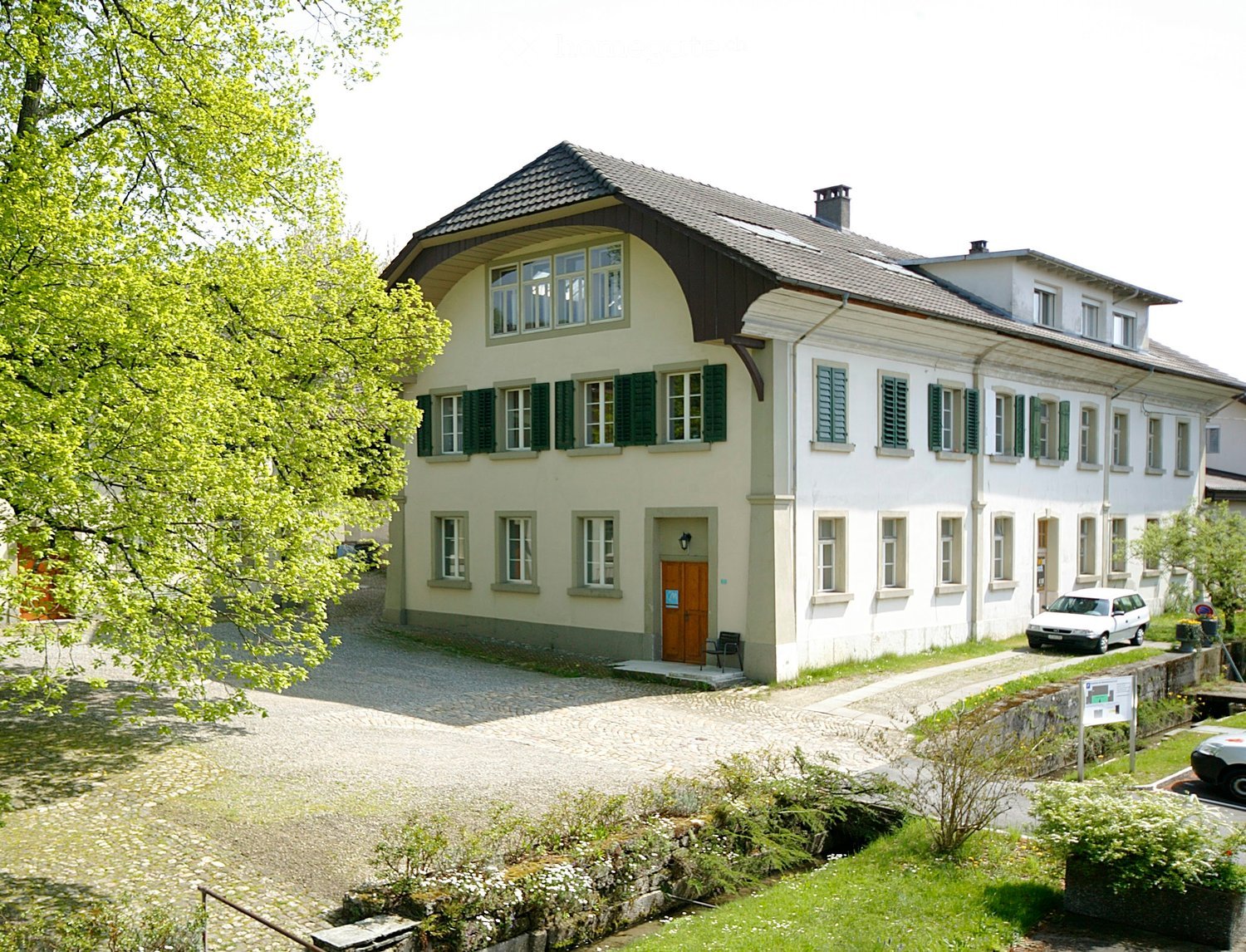Two-story house with light yellow and white exterior, green shutters, multiple windows, and a gravel driveway.