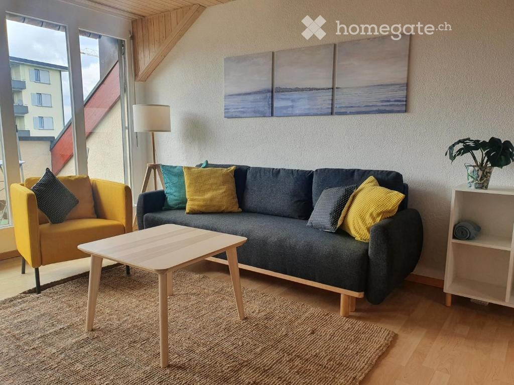 Modern living room with large windows offering a view, featuring a grey sofa with colorful pillows, a yellow armchair, a wooden coffee table, a floor lamp, a shelf with a plant, and abstract artwork on the wall.