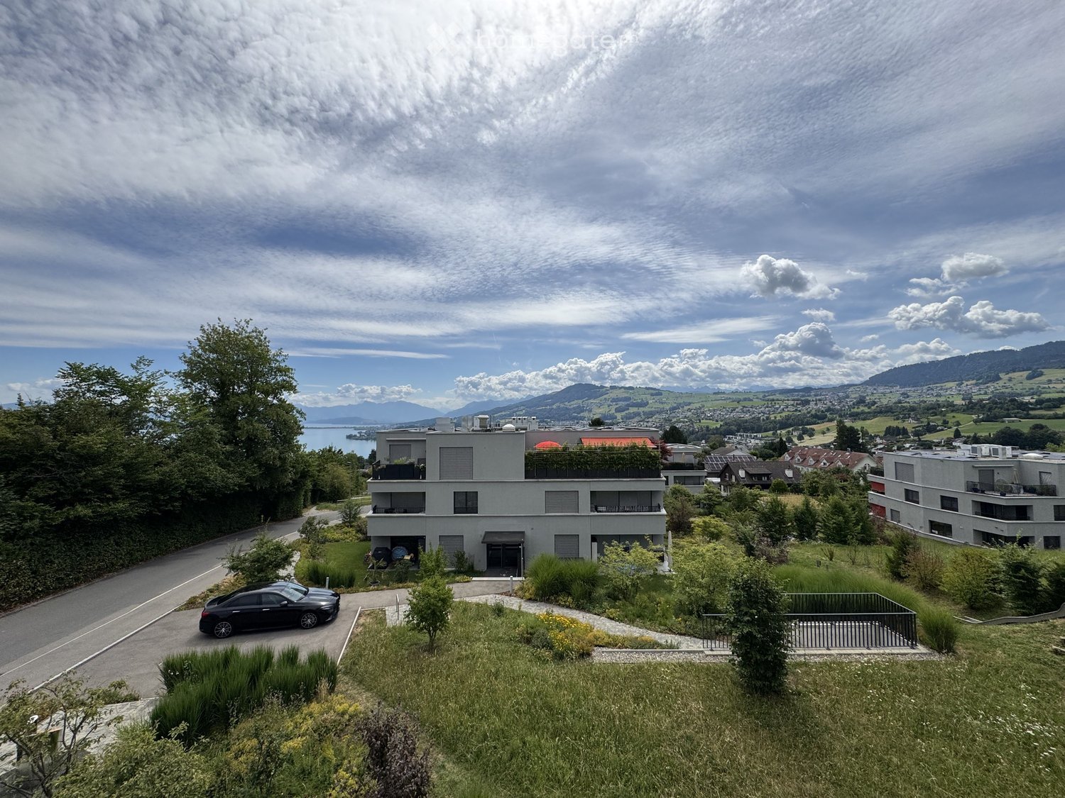 Modern two-story house with a flat roof, large windows, and a balcony, situated on a hillside with a scenic view of a lake and mountains in the background.