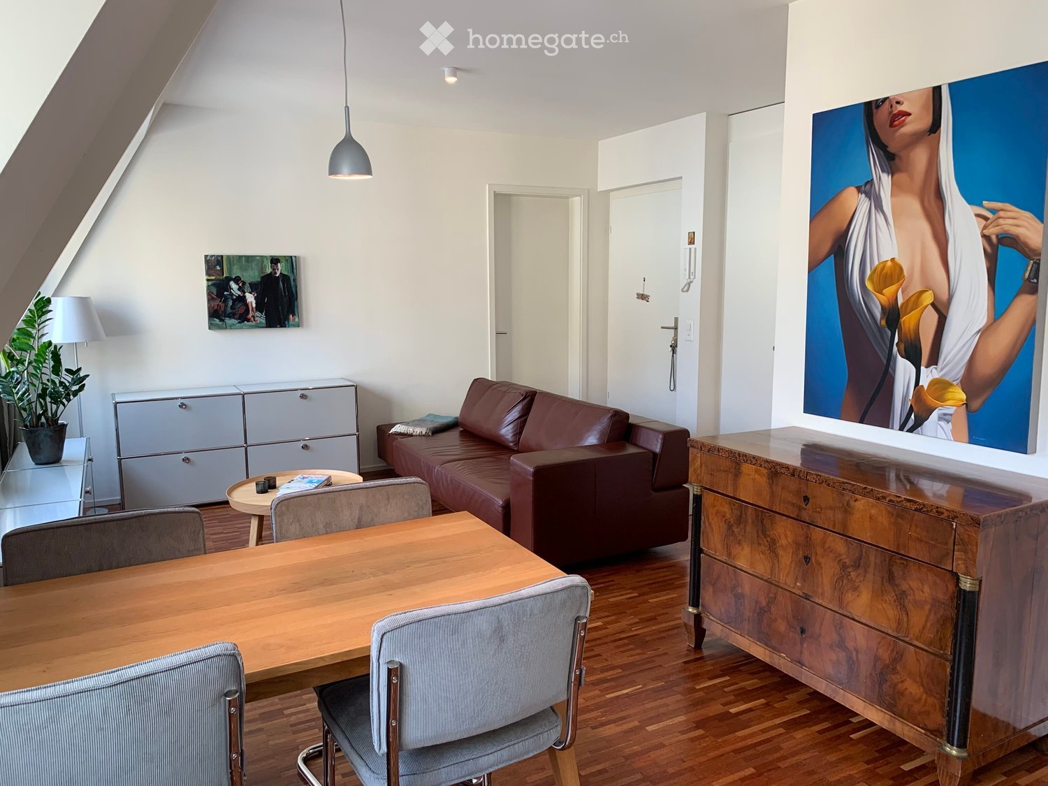 Modern living room with wooden flooring, a brown leather sofa, a wooden dining table with chairs, a side table, a cabinet, a lamp, and artwork on the walls.