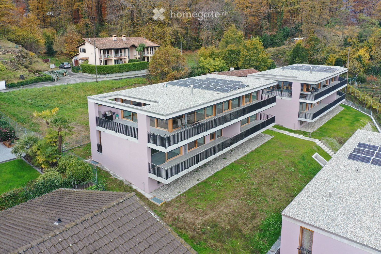 Aerial view of a modern, multi-story residential building with pink exterior, large windows, and multiple balconies. The roof features solar panels, and the surrounding area includes well-maintained lawns, trees, and neighboring houses.