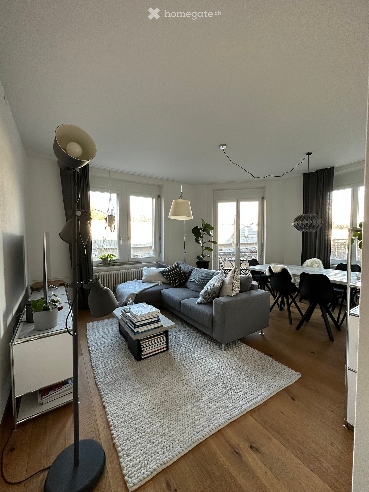 Modern living room with a grey sectional sofa, coffee table with books, white shag rug, wooden floor, large windows, and pendant lights.