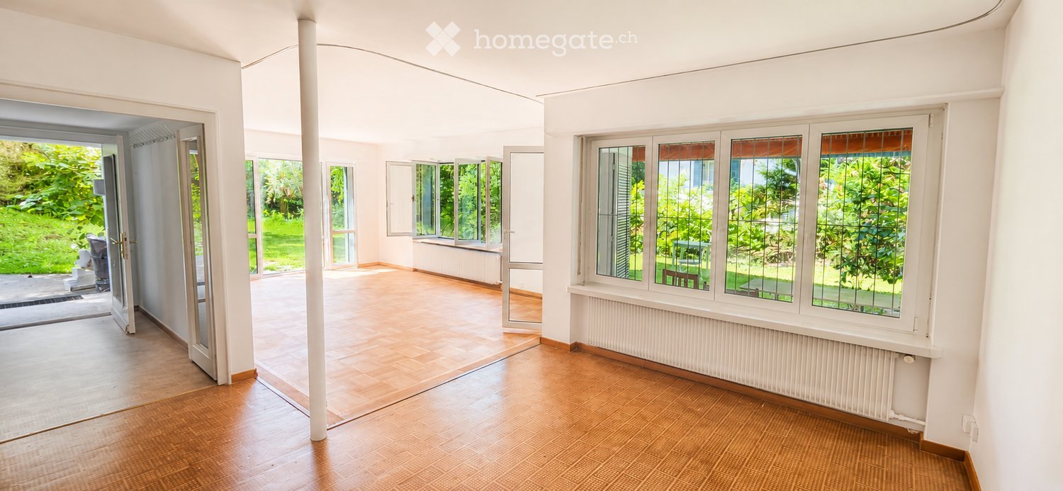 Bright, open room with large windows and wooden flooring, leading to a garden area.