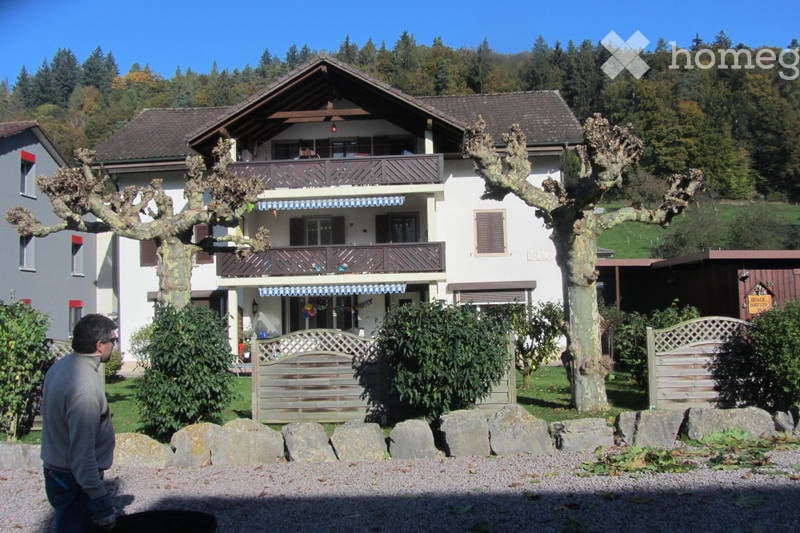 A two-story house with a balcony, surrounded by trees and greenery. The house has a traditional design with wooden elements and a stone pathway leading to the entrance.