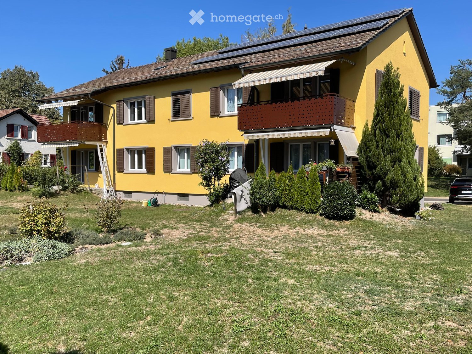 Two-story yellow house with brown shutters, multiple windows, balconies, and solar panels on the roof. The front yard has a well-maintained lawn, shrubs, and a parked car.