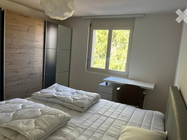 Modern bedroom with a large bed, white bedding, a desk with a chair, and a wardrobe. The room features a large window with a view of greenery outside and a contemporary ceiling light fixture.