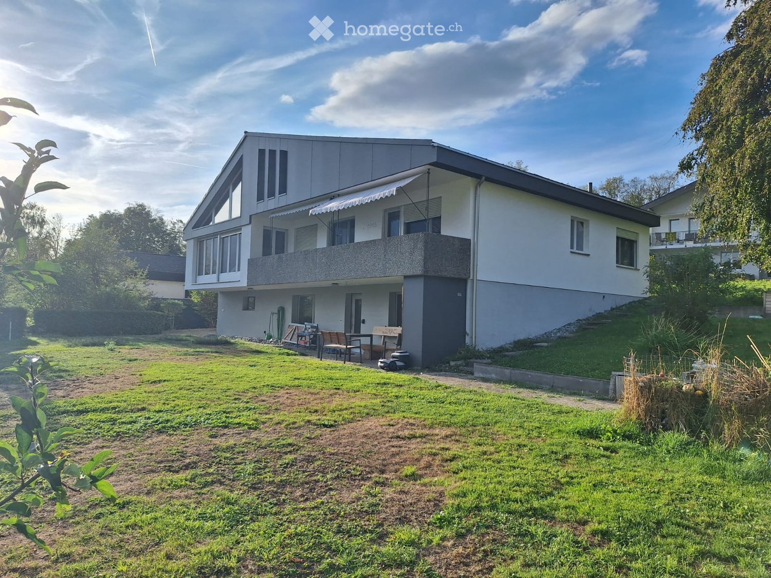 Two-story modern house with white exterior, large windows, and a balcony on the upper level. The house is surrounded by a well-maintained lawn and garden area.