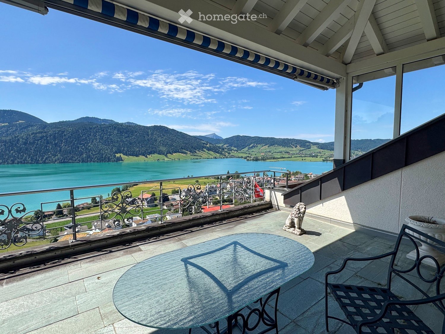 Balcony with a scenic view of a lake and mountains, featuring a glass table, a metal chair, and a decorative statue.
