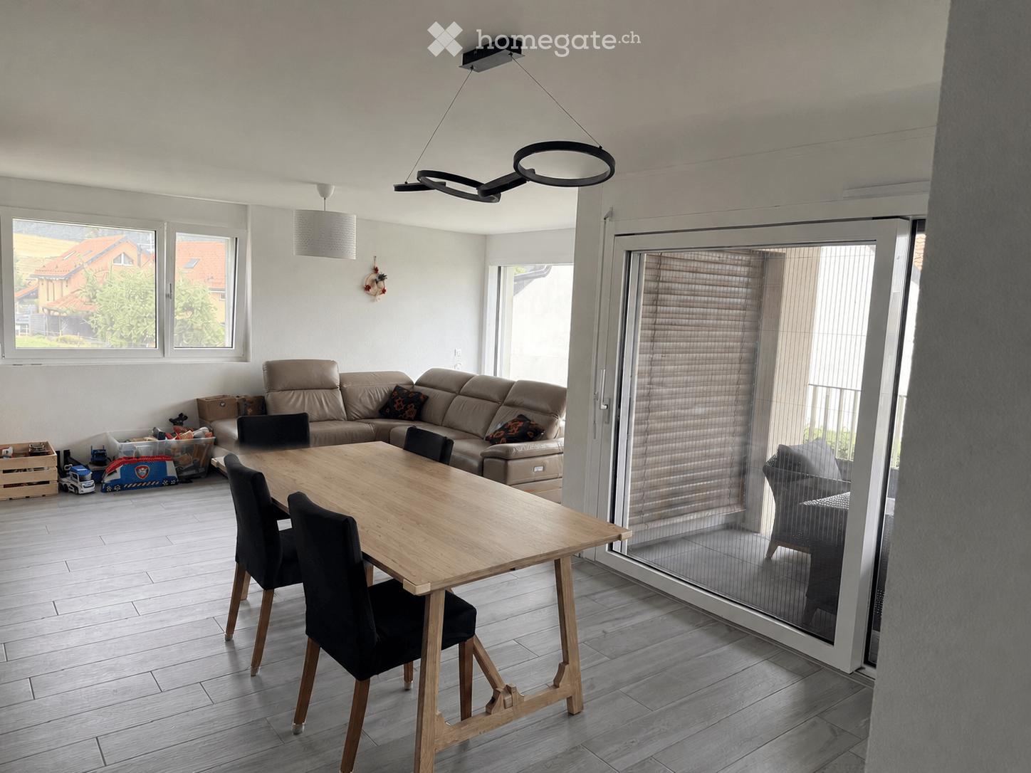 Bright living and dining area with a beige sofa, wooden dining table, black chairs, and a large window with blinds.