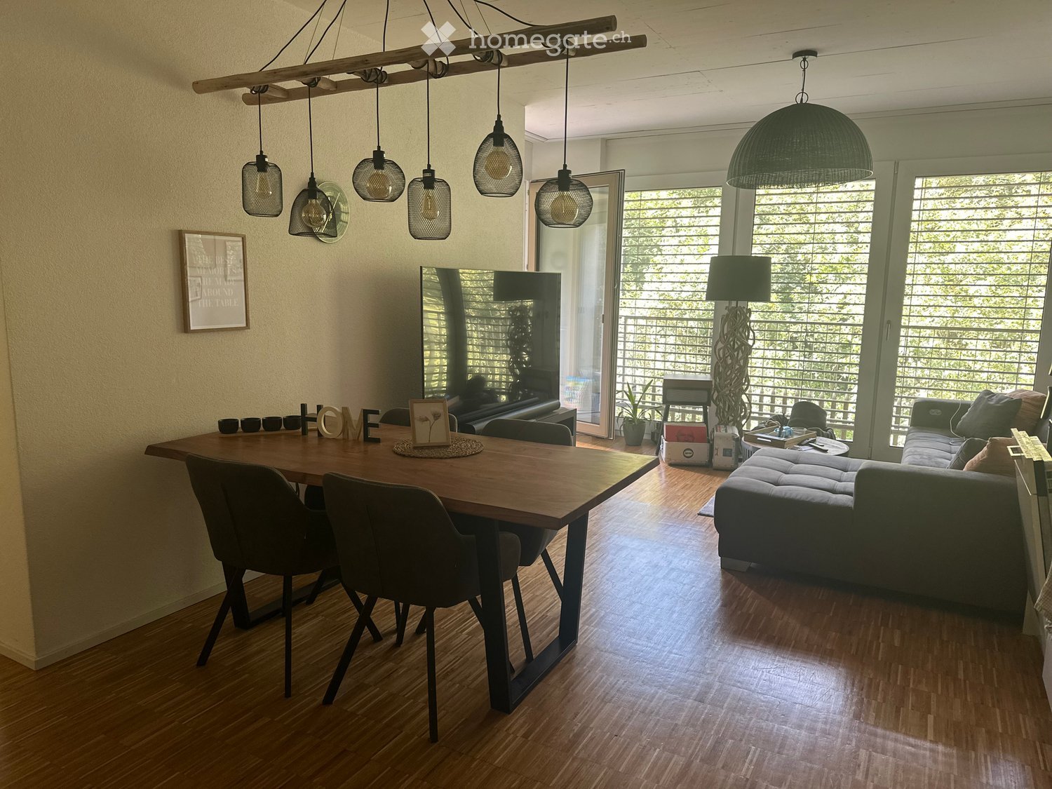 Modern living and dining area with hardwood floors, a large sofa, a dining table with chairs, pendant lights, and a large mirror. The space features large windows with blinds and a view of greenery outside.