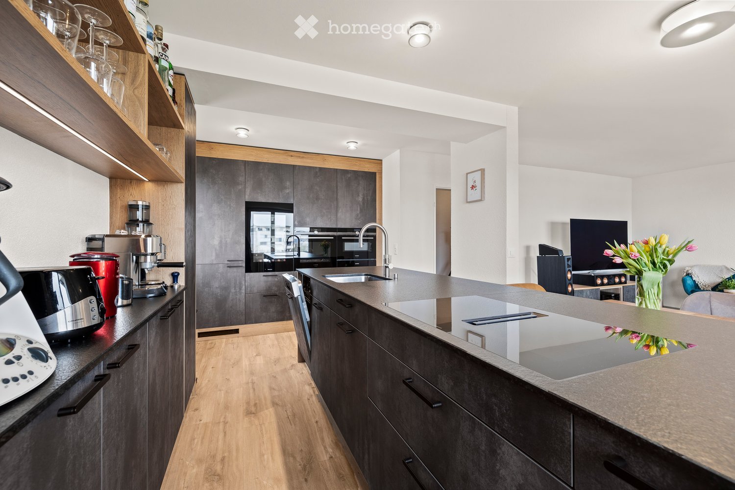 Modern kitchen with dark grey cabinets, wooden shelves, stainless steel appliances, and a central island with a built-in induction cooktop. The space features a coffee maker, toaster, and a vase with flowers on the island.
