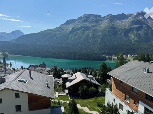 Panoramic view of a town nestled in a valley surrounded by mountains, with a lake in the foreground and a mix of residential and historic buildings.