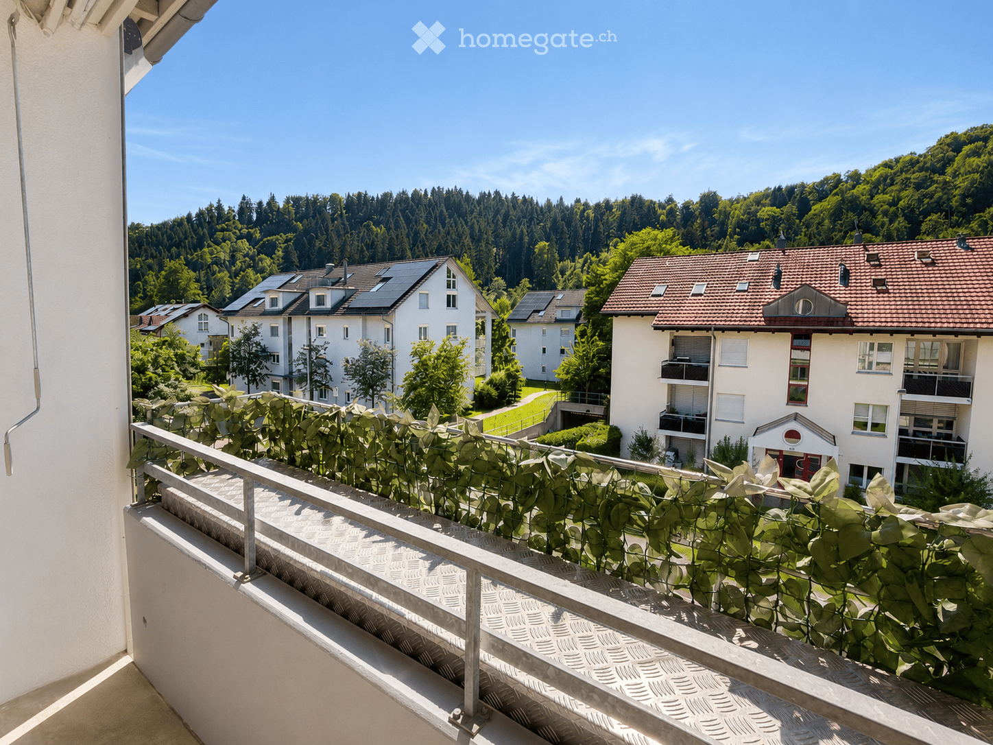 Balcony with metal railing, lush green plants, overlooking a residential area with houses, trees, and a clear blue sky.