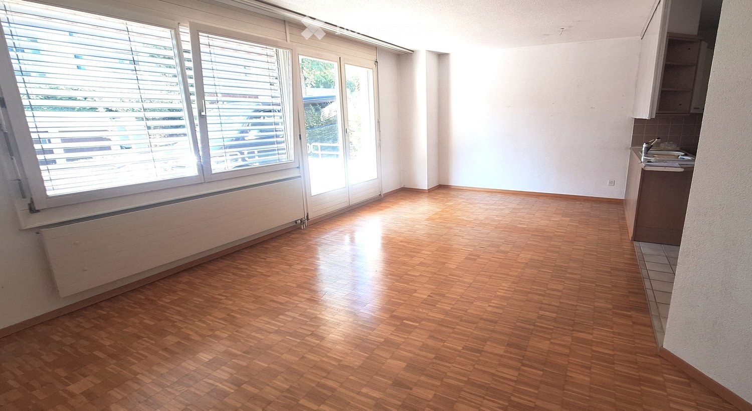 Empty room with wooden flooring, large windows with blinds, and a small kitchen area with a sink and cabinets.