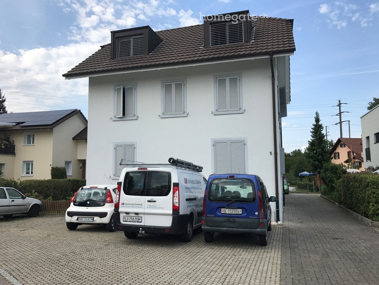 Two-story white house with multiple windows and a tiled roof, with a paved driveway and several parked vehicles including a van and a car.