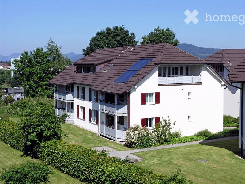 A modern two-story residential building with white walls, red shutters, and a brown tiled roof. Features include multiple balconies, a solar panel on the roof, and a well-maintained lawn with hedges and trees in the foreground.