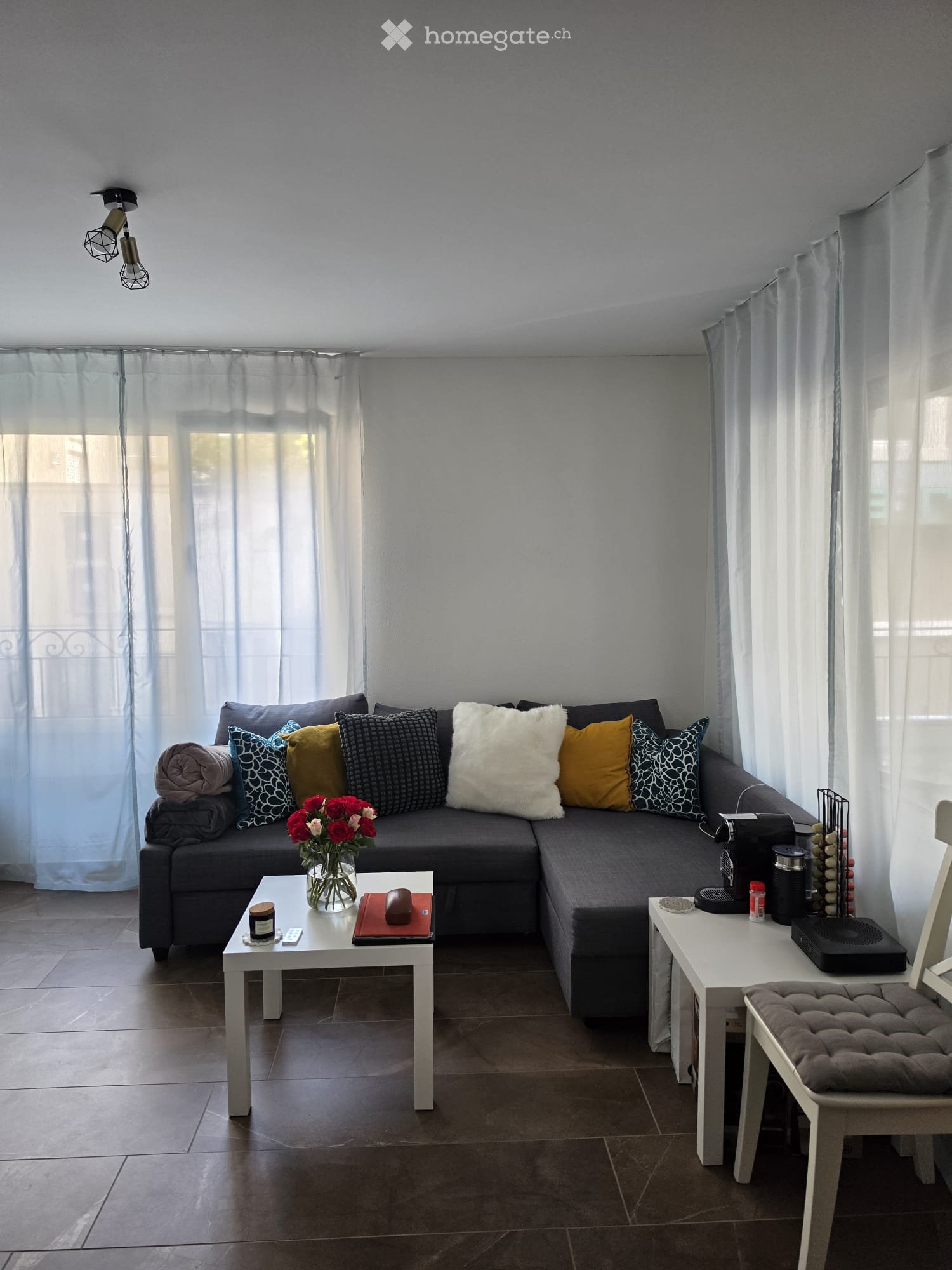 Modern living room with grey sectional sofa, colorful throw pillows, coffee table with vase of red flowers, remote control, book, and candle. There is a white side table with a laptop, cup, and other small items. The room features large windows with sheer