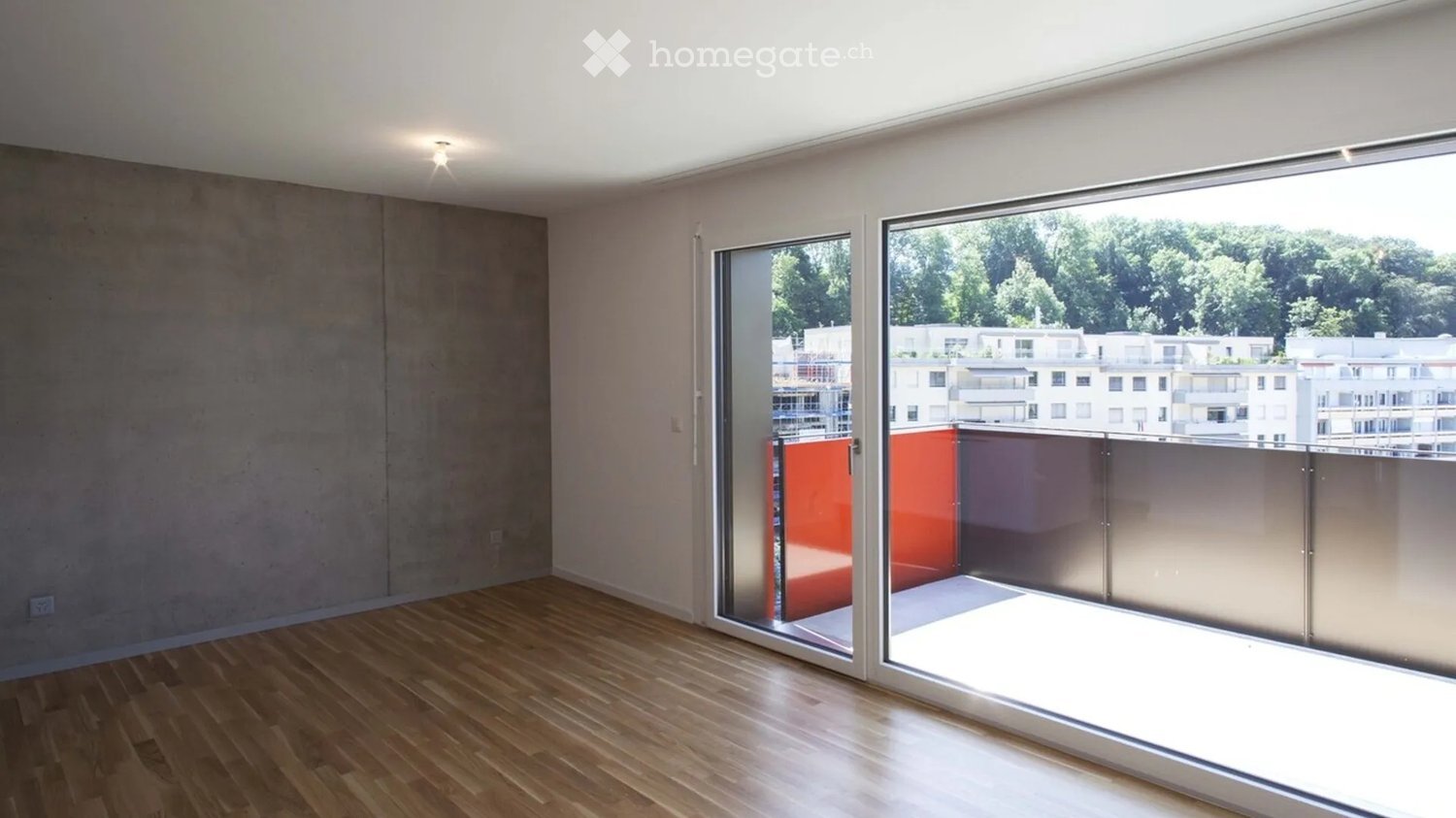 Empty room with wooden flooring, large sliding glass doors leading to a balcony, and a view of surrounding buildings and greenery.
