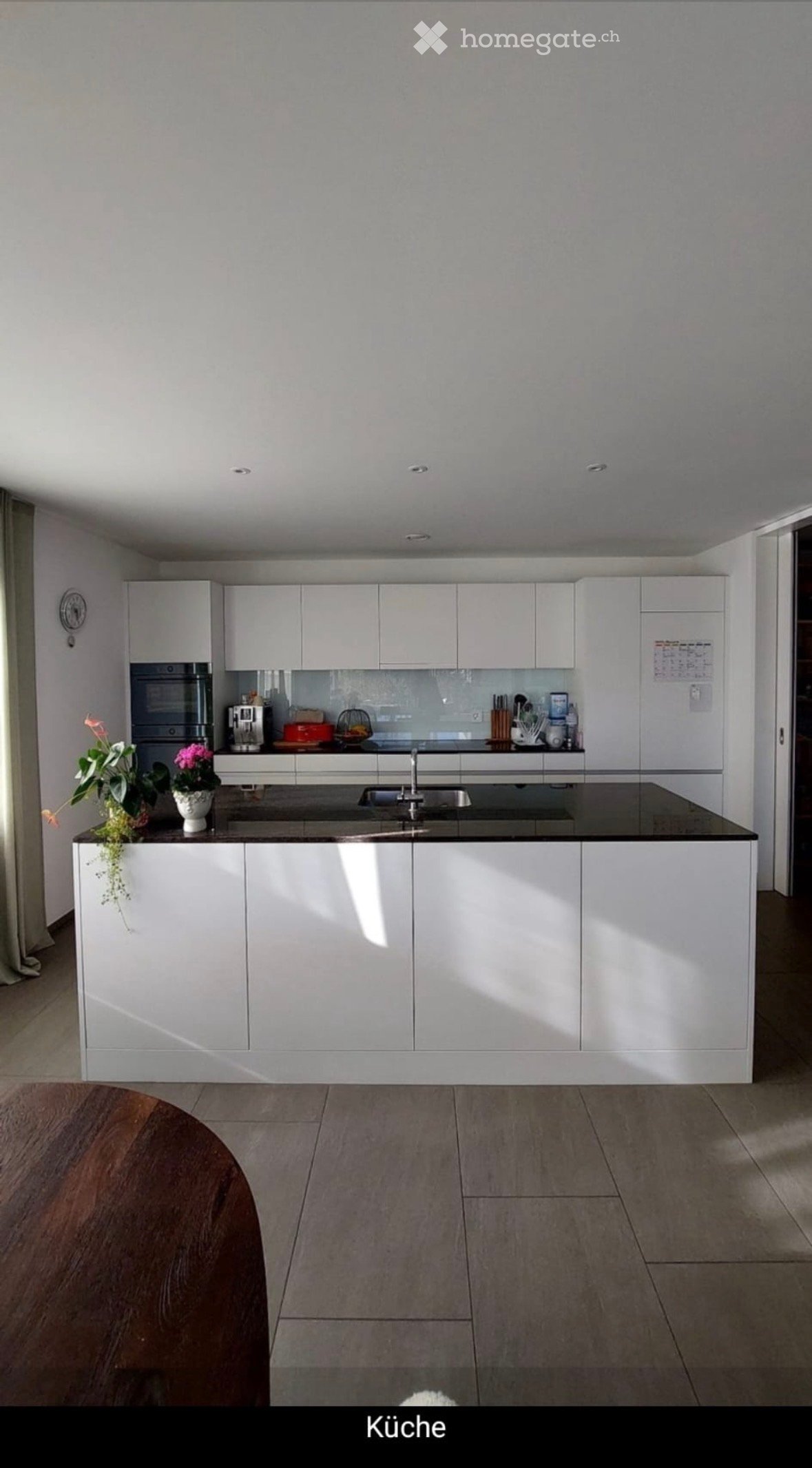 Modern kitchen with white cabinets, a central island, and a black countertop. Features include a sink, faucet, oven, coffee maker, and decorative flower pots on the island.
