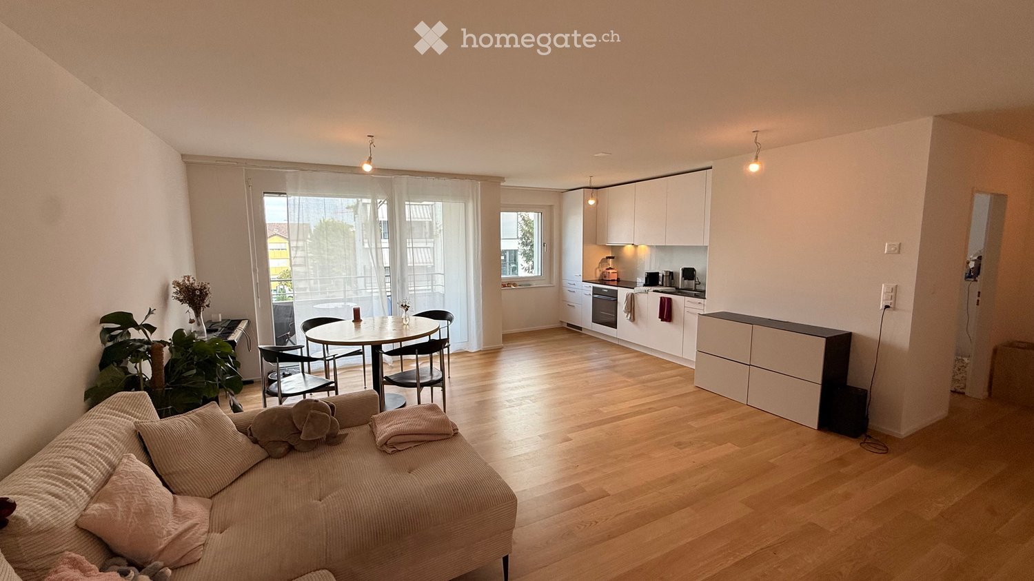 A modern, open-plan living and dining area with a sofa, dining table, chairs, and a fitted kitchen with white cabinets and appliances.