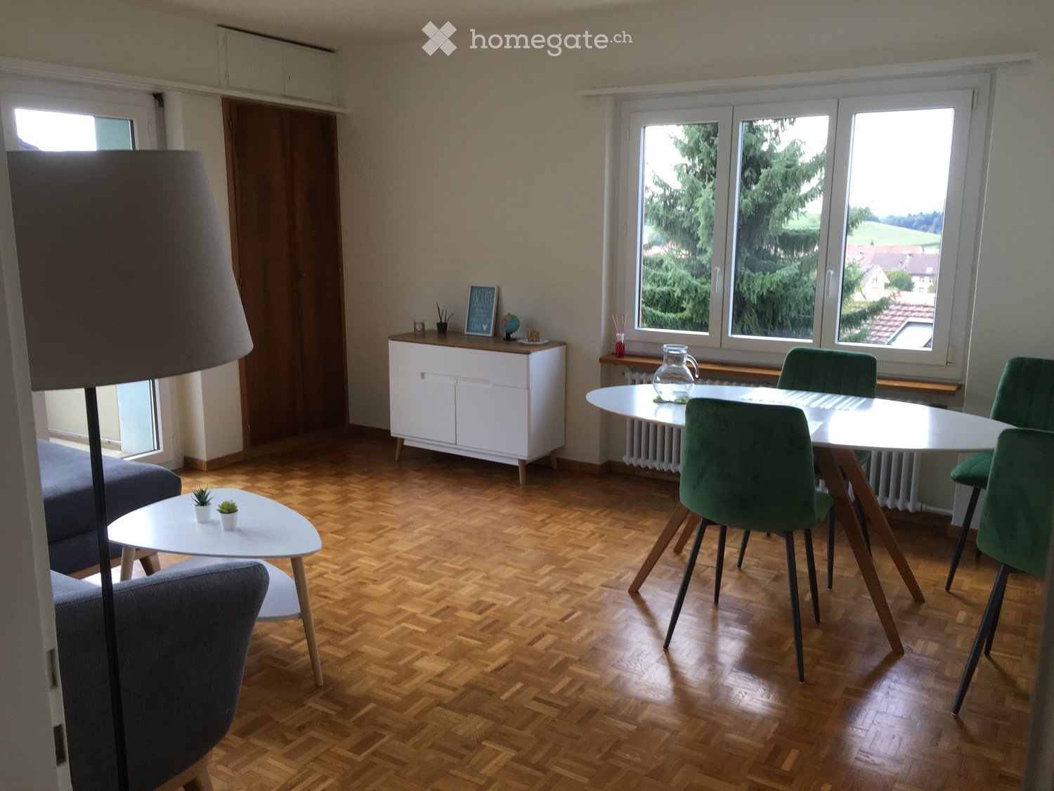 Bright living and dining area with wooden flooring, large windows, a grey sofa, a white coffee table, a dining table with four green chairs, a white cabinet, and a floor lamp.