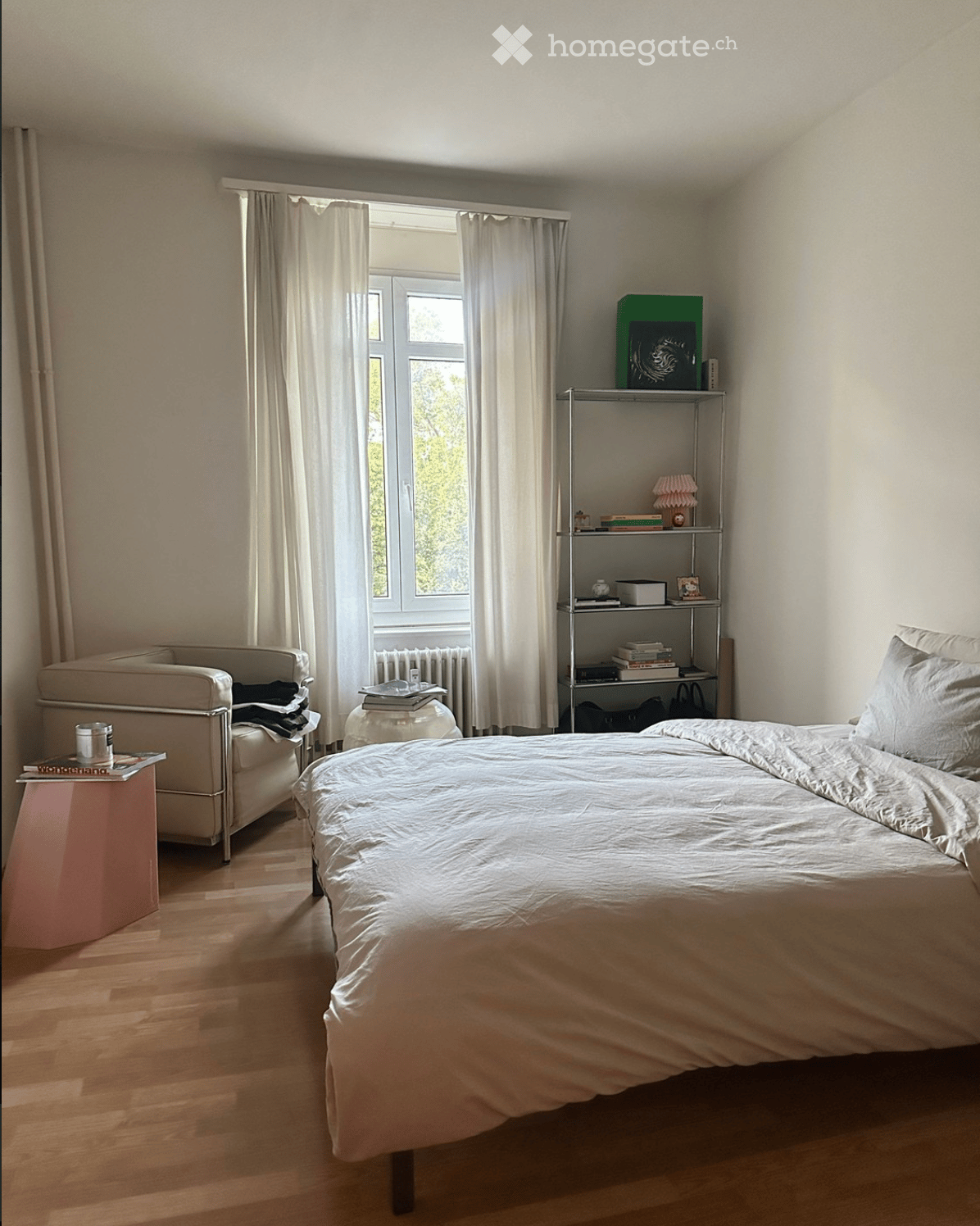A well-lit bedroom with a large bed, a small sofa, a side table, and a metal shelf with books and decorative items. The room has wooden flooring and large windows with white curtains.