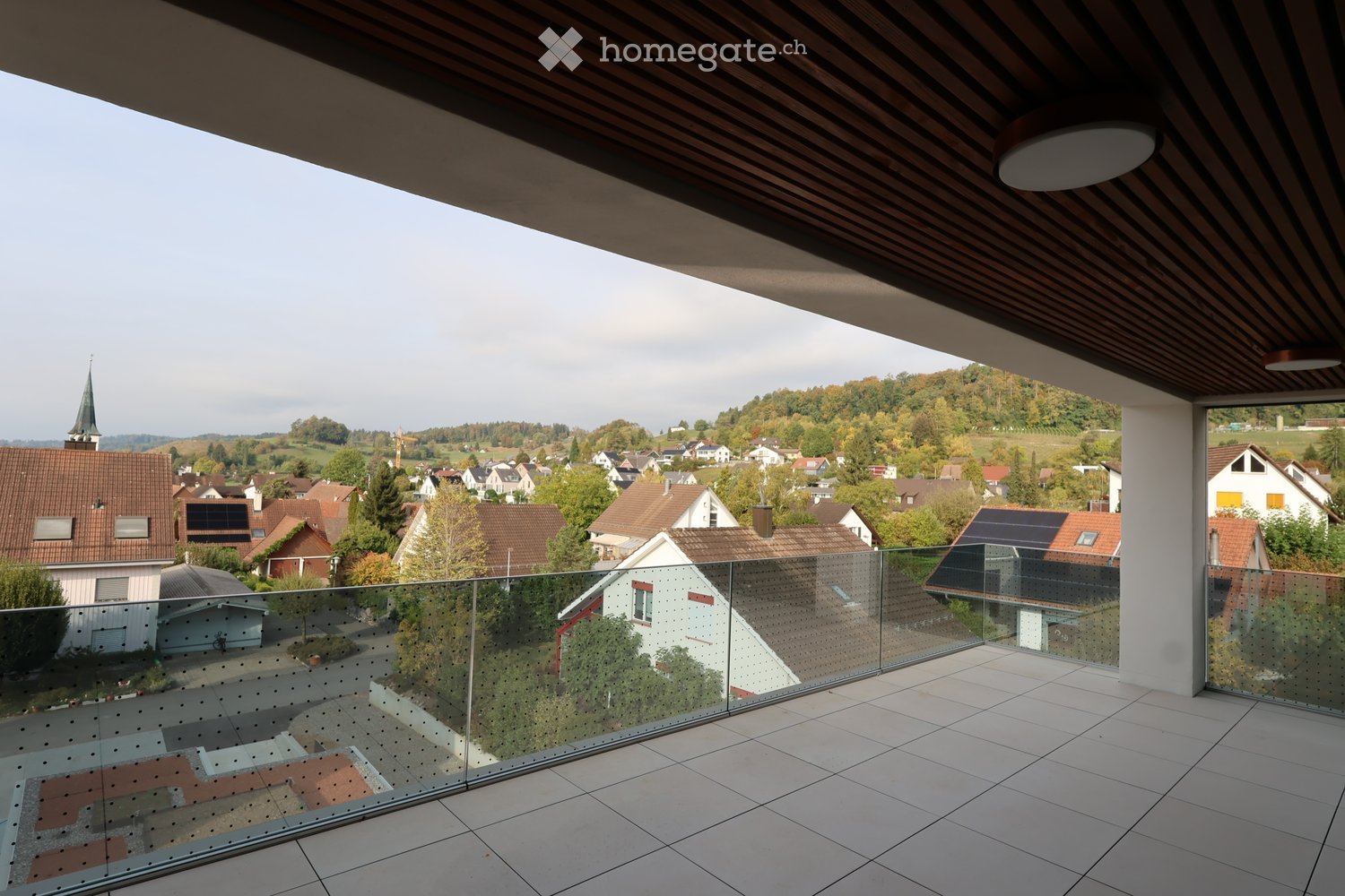 Modern balcony with glass railing, tiled floor, and wooden ceiling, overlooking a town with houses and a church spire.