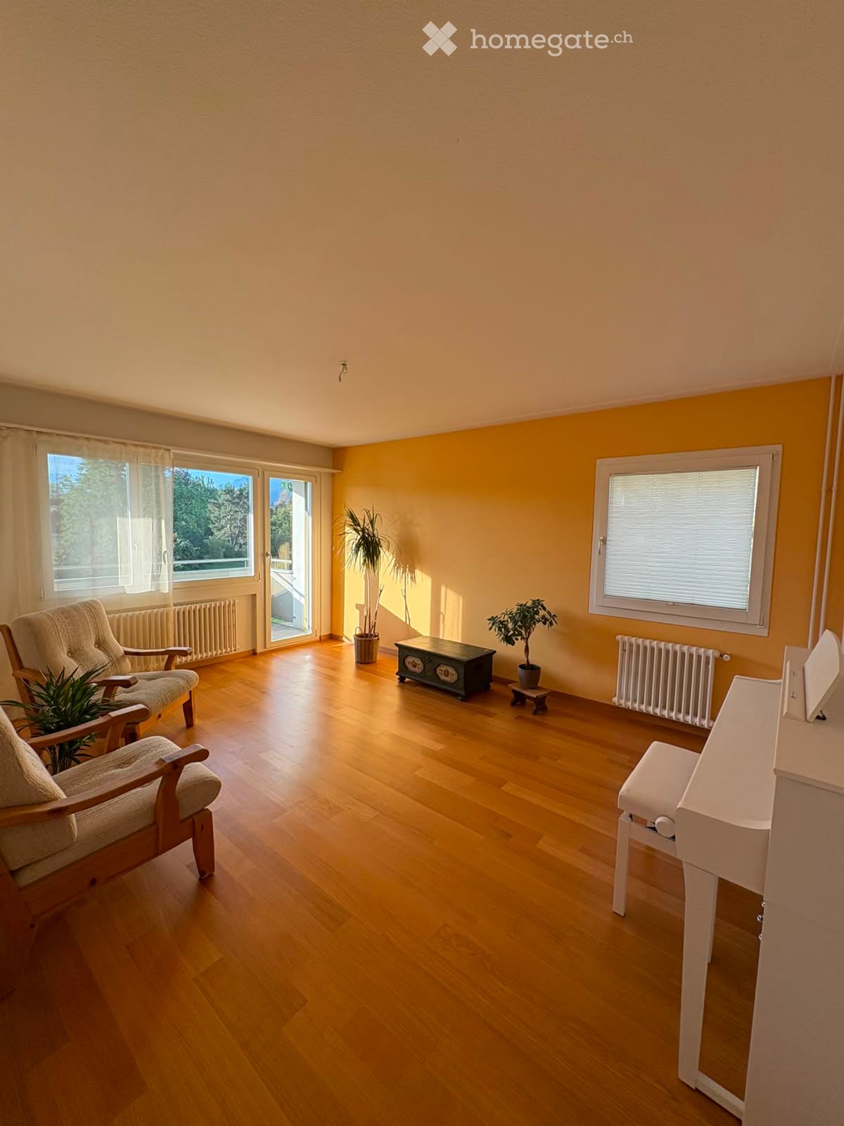 Bright living room with wooden flooring, large windows, and a sliding glass door leading to a balcony. Features include a sofa, a small table, two armchairs, and potted plants.