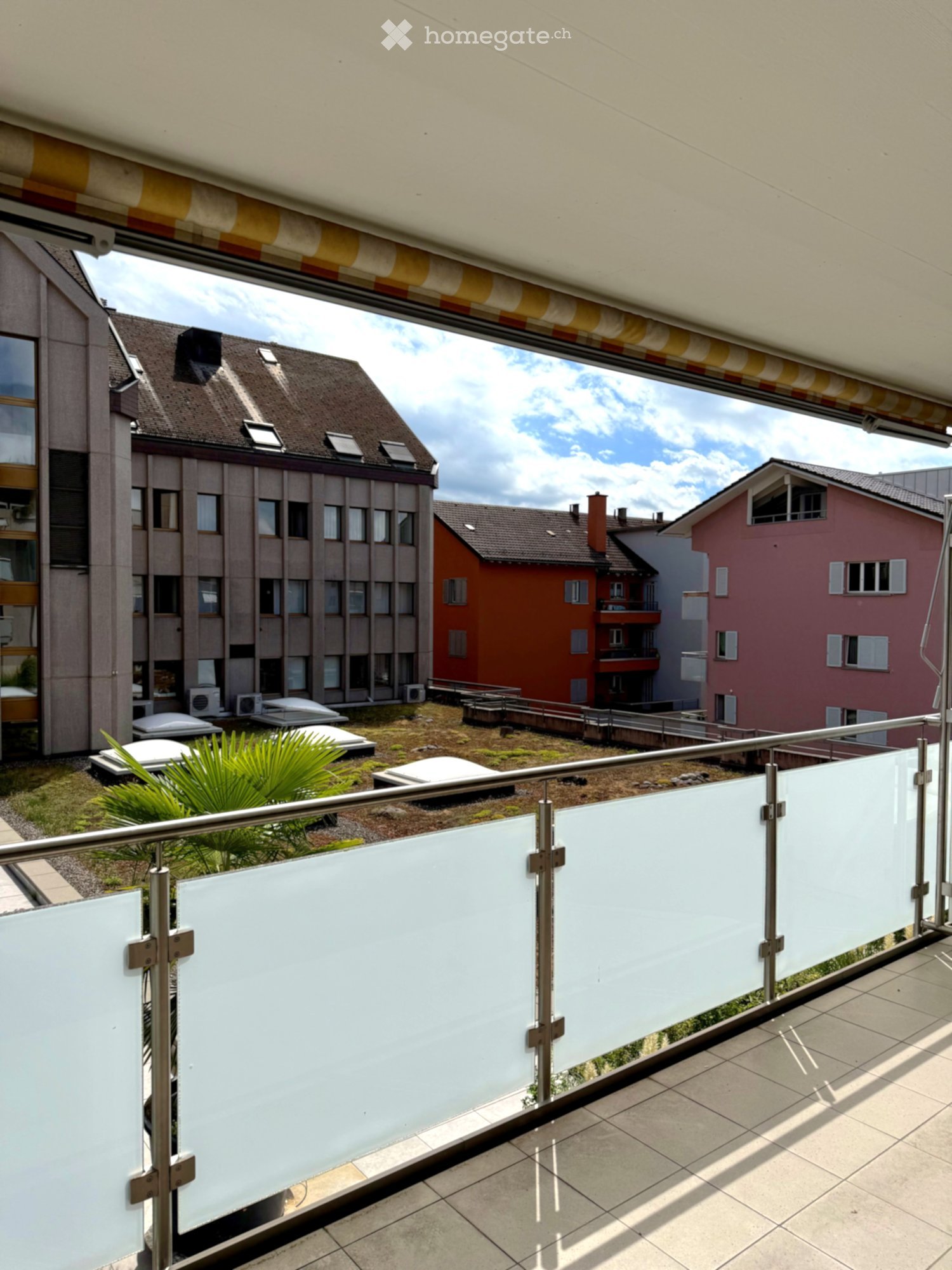 Balcony with glass railing, tiled floor, and view of surrounding buildings and greenery.