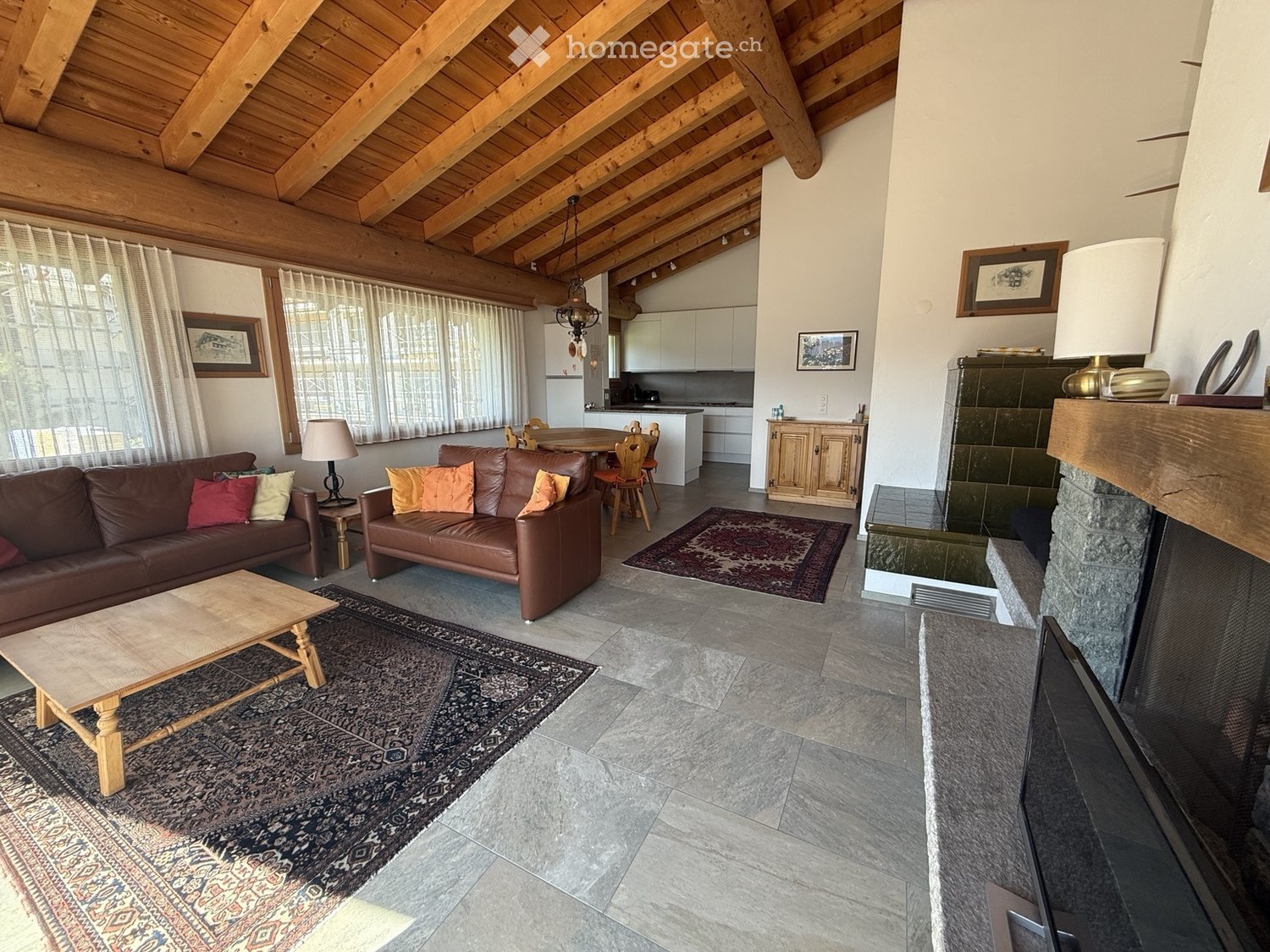 A spacious living room with exposed wooden beams, a stone fireplace, two leather sofas, a coffee table, a dining area with a table and chairs, and a modern kitchen in the background.