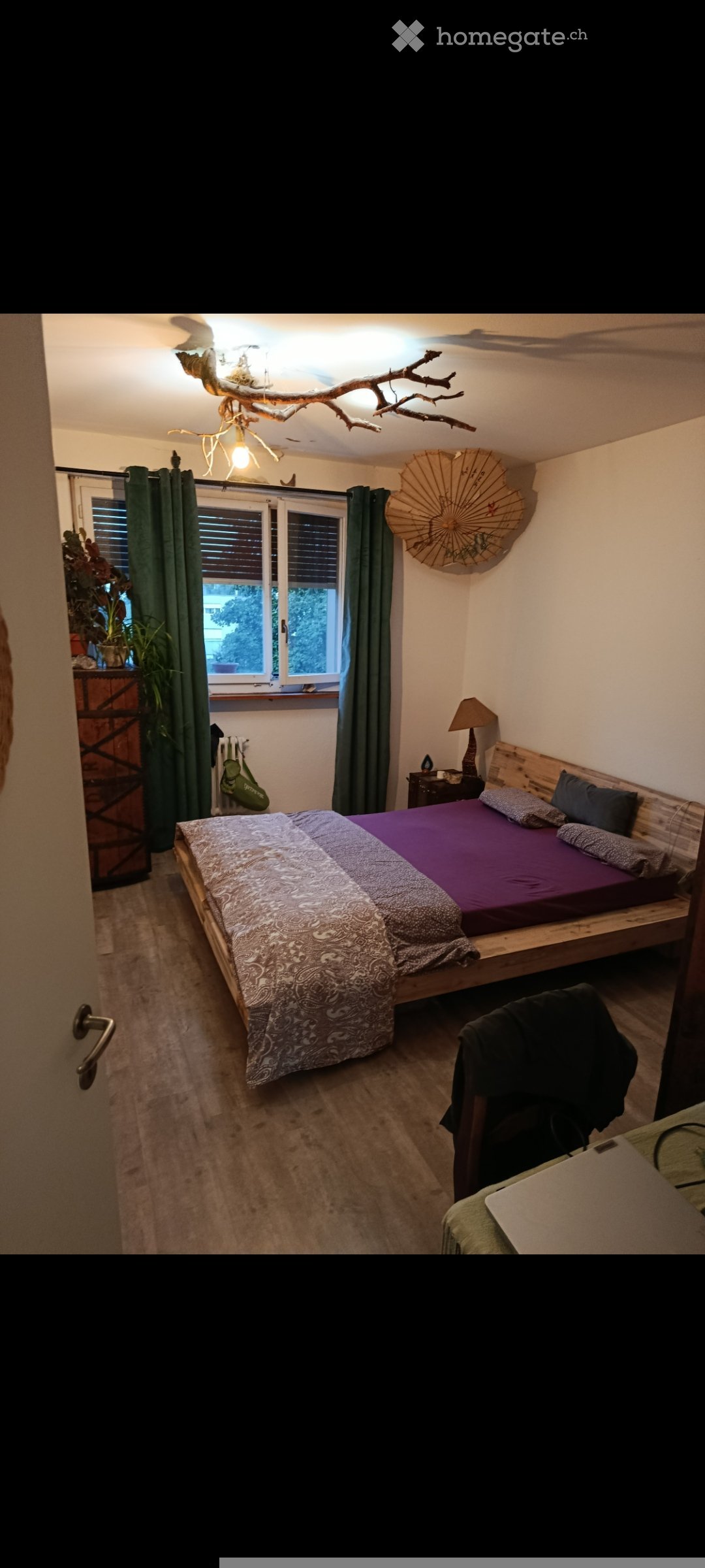 Cozy bedroom with a wooden bed, purple bedding, nightstand with lamp, green curtains, and a decorative umbrella on the wall.