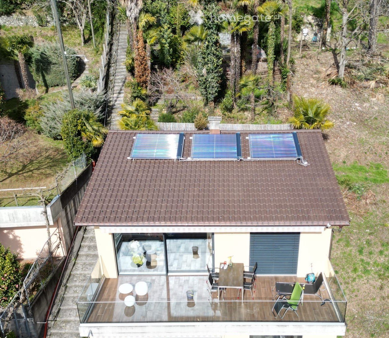 A two-story house with a tiled roof, solar panels, and a spacious balcony with outdoor furniture including a table, chairs, and a lounge chair.