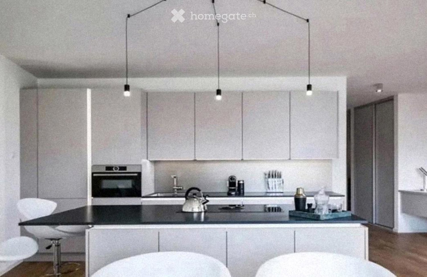 Modern kitchen with white cabinets, black countertop, built-in oven, and pendant lights. Includes stools, teapot, coffee maker, and various small items on the counter.