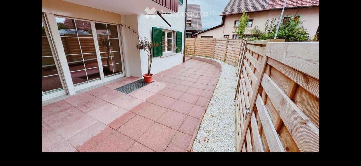 A paved patio area with pink tiles, a small potted plant, and a wooden fence surrounding the space. The patio is adjacent to a house with large glass doors and green shutters.