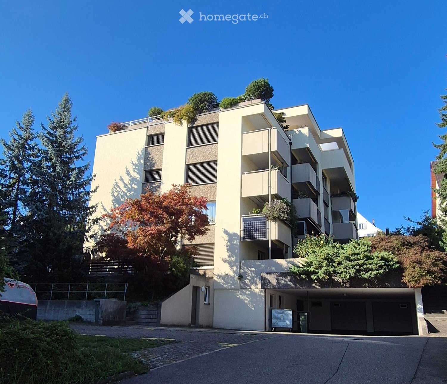 Modern multi-story residential building with balconies, greenery on the roof, and a covered parking area at the base.