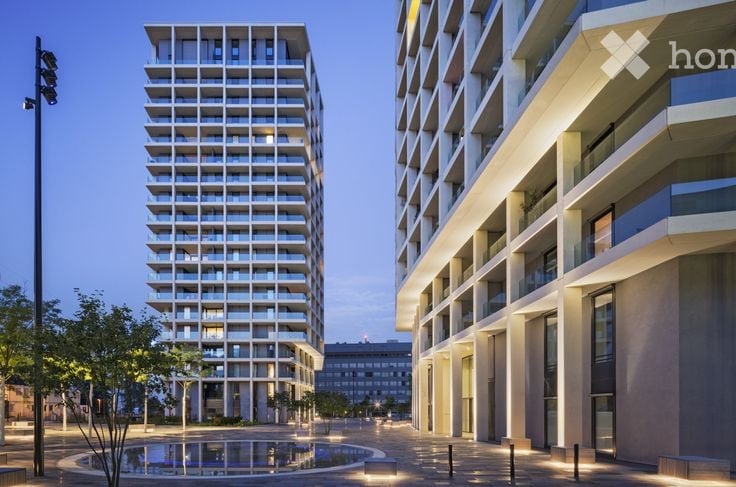 Modern high-rise residential buildings with clean lines and large windows, surrounded by a paved plaza with a small water feature and trees.